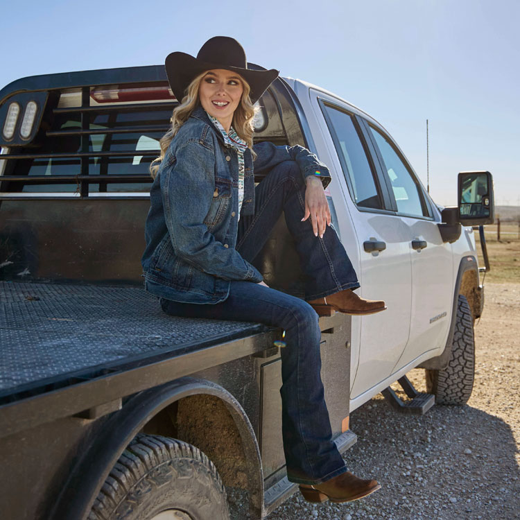 A woman wearing women’s Annie Up 13” Western boot in tan and Roper Felt Western Hat in black sitting on a flatbed truck.